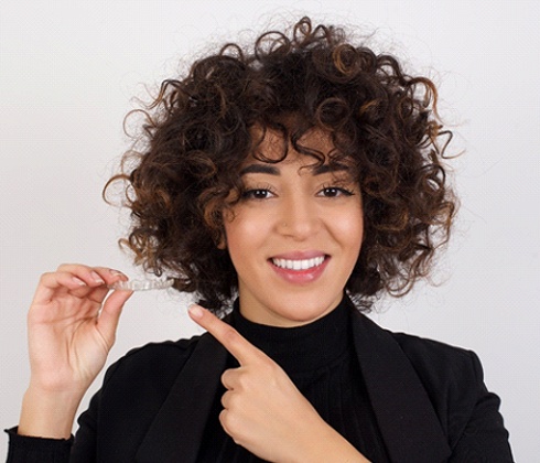 A woman with curly hair holding a clear aligner while pointing to it and smiling in Cleburne
